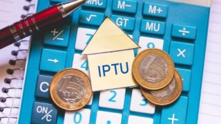 IPTU: imagem mostra uma calculadora em cima de uma caderno. Em cima da calculadora há uma caneta, três moedas de um real e um recorte de papel em formato de casa, com a sigla IPTU escrita dentro.