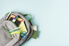Uma mochila está aberta e possui diversos materiais escolares, como canetas, tesoura e caderno.