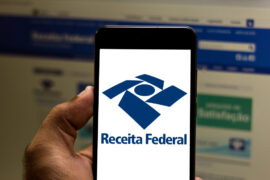 Imposto de Renda: um homem segura um celular e, na tela, é mostrado o logo da Receita Federal. Atrás o notebook está com o site do órgão aberto.