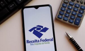 Imposto de Renda: imagem mostra um celular com o logo do aplicativo IRPF, da Receita Federal. Ao lado há uma caneta e uma calculadora