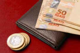 Consórcio de serviços: algumas notas de cinquentas reais estão colocadas em cima de uma carteira de couro marrom. Ao lado ha moedas de um real empilhadas.