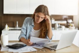 Empréstimo para quitar dívidas: uma mulher está sentada à mesa, enquanto lê documentos. Há, na mesa, outros papéis, um notebook, uma caneca e uma calculadora.