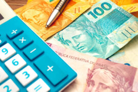 Desenrola Brasil: algumas notas de dinheiro estão espalhadas. Em cima há uma caneta e uma calculadora azul com branco.