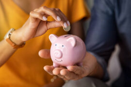 Poupança: uma mulher insere uma moeda em um cofre em formato de porco, enquanto um homem segura o objeto.