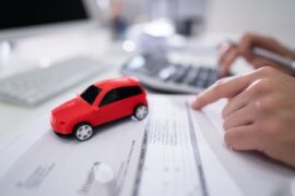 Taxas de refinanciamento de veículo: uma pessoa observa alguns dados contidos em uma folha, enquanto mexe na calculadora. Ao lado há uma miniatura de automóvel e um computador.