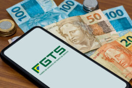 Consórcio de imóvel: celular mostra o logo do FGTS e, ao lado, há notas de cinquenta e cem reais, além de moedas.